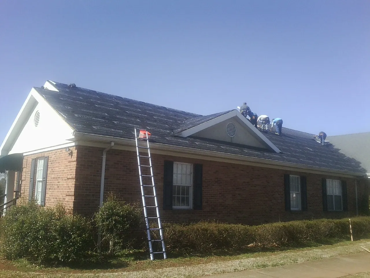 Crew tearing shingles off a brick ranch roof with a ladder set — Emergency Roof Repair work by our crew in Sealy