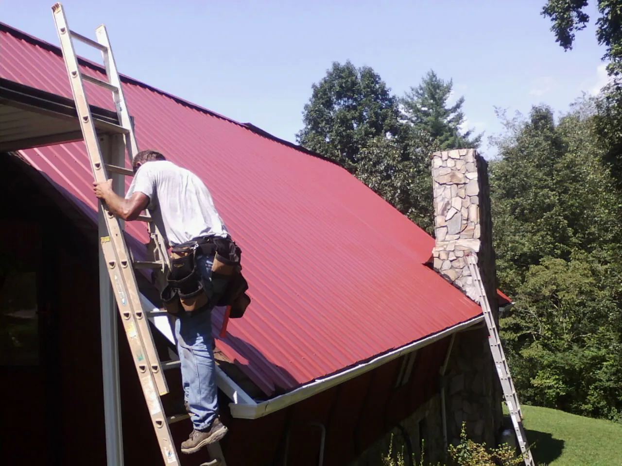 Roofer on a ladder beside a red metal roof and stone chimney — Soffit & Fascia Repair by Roofing Brothers in Sealy